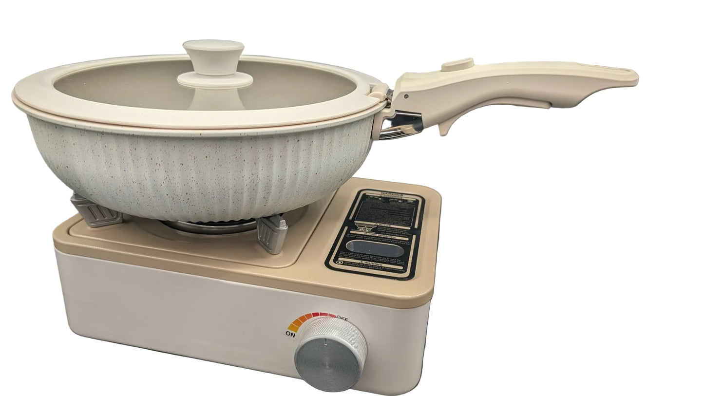 A ceramic non-stick pan with a matching lid sits on a compact portable electric burner. The pan has a ribbed off-white exterior and a cream-colored handle, while the burner is beige with a black control panel and a silver dial, set against a dark gray background.
